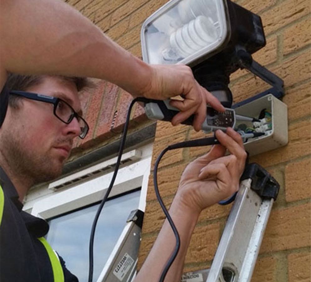 Electrician on a ladder wiring an outdoor floodlight and sensor to brickwork