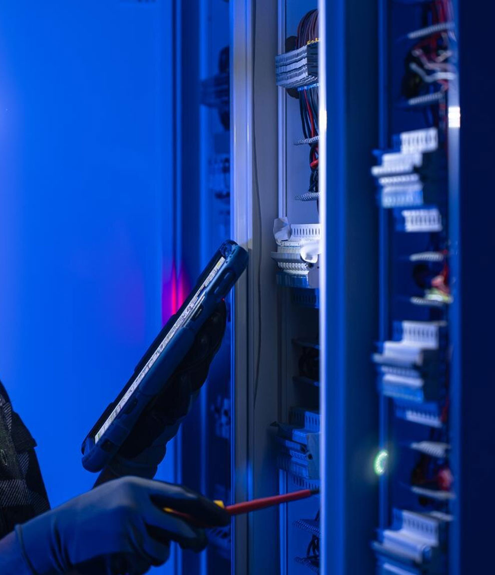 Engineer holding tablet and screwdriver beside tall industrial switchgear cabinets