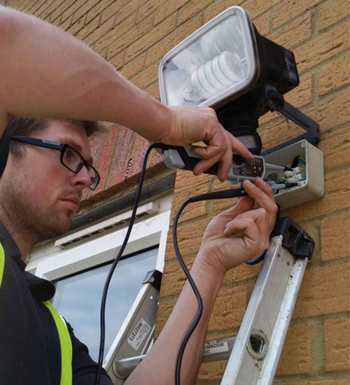 Man on a ladder wiring an outdoor floodlight on a brick wall