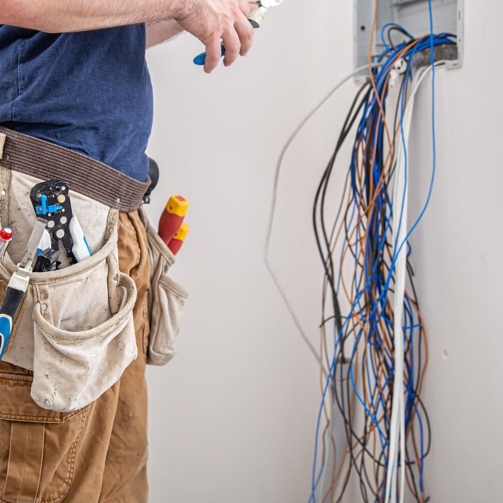 Electrician in a tool belt beside loose cables hanging from a wall enclosure